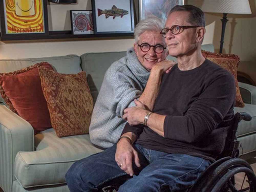 man in wheelchair and wife sitting next to him on couch