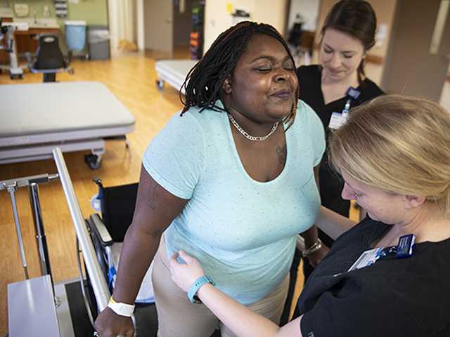 Two healthcare providers helping woman standup from wheelchair.